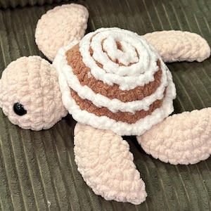 Puede incluir: Una tortuga de peluche hecha a mano con un caparazón en forma de rollo de canela. El caparazón es marrón y blanco, y el cuerpo y las extremidades de la tortuga son de un rosa suave y claro. La tortuga tiene un ojo negro.