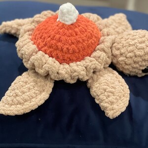 以下が含まれることがあります： 赤と白の殻を持つ、かぎ針編みで作ったウミガメのぬいぐるみ。青い表面の上に横たわっています。