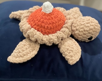 May include: A crocheted sea turtle toy with a red and white shell. The turtle is lying on a blue surface.