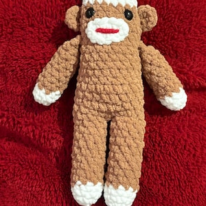 May include: A handmade sock monkey doll with a brown body, white accents on the face, hands, and feet, and a red pom-pom on top of its white head. The doll has black eyes and a red mouth. The plush toy is lying on a red, fuzzy surface.