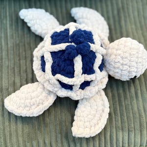 May include: A handmade plush turtle in white and navy blue. The turtle has a textured, fluffy appearance, with a white shell outline and flippers. The turtle's shell is navy blue with white accents. The head is a round, white ball.