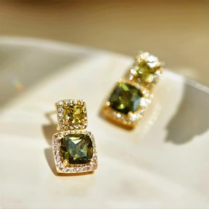 May include: A pair of gold earrings featuring green and clear gemstones. The earrings have a square design with a smaller square gemstone on top of a larger square gemstone.
