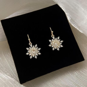 May include: A pair of gold snowflake earrings with clear crystal accents. The earrings are on a black velvet background.
