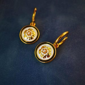 May include: A pair of gold hoop earrings with white and black enamel flower accents. Each flower has a small pearl in the centre.