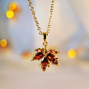 May include: A gold chain necklace with a pendant shaped like a maple leaf. The leaf is adorned with red gemstones.