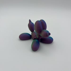 Puede incluir: Un pequeño animal de globo de color morado iridiscente con forma de perro. El animal de globo está hecho de un material brillante y metálico.