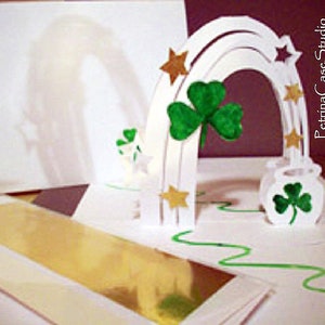 May include: A white paper pop-up card with a rainbow arch, shamrocks, and gold stars. The card is open and the inside is decorated with a gold foil strip and a green wavy line.