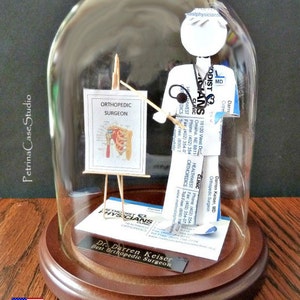May include: A glass dome with a wooden base displays a paper figure of an orthopedic surgeon holding a stethoscope and a pointer. The figure is standing in front of a small easel with a diagram of a shoulder joint. The base has a plaque that reads "Dr. Darren Keiser Best Orthopedic Surgeon".