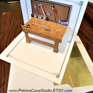 May include: A pop-up card with a 3D image of a workbench with tools. The workbench is made of wood and has a pegboard with tools hanging on it. The card is white with a gold border and the text "www.PetrinaCaseStudio.ETSY.com NO. 954" on the bottom.