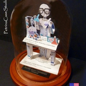 May include: A papercraft figurine of a chemist wearing a lab coat and glasses, holding a beaker and test tube. The figurine is displayed on a table with a business card that reads "Burton Chemist" and "Research Laboratories". The figurine is encased in a glass dome on a wooden base.