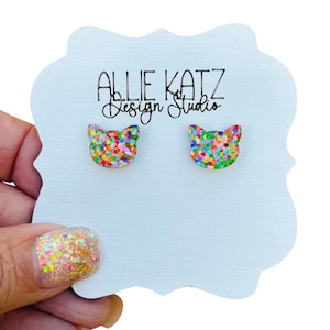 May include: A pair of colorful cat-shaped stud earrings with a rainbow glitter finish. The earrings are displayed on a white card with the text "Allie Katz Design Studio".