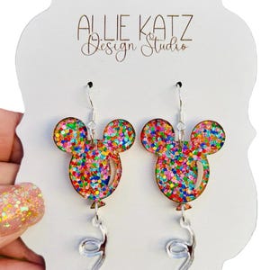 May include: A pair of dangle earrings featuring a colorful confetti balloon design with a silver ribbon. The balloon is shaped like Mickey Mouse ears. The earrings are on a white card with the text "Allie Katz Design Studio".