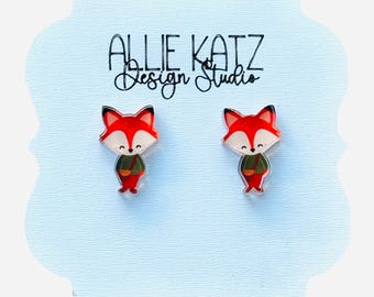 May include: A pair of fox-shaped earrings. The earrings feature a red fox with a white face, black ears, and a green jacket. The earrings are on a light blue card with the text "ALLIE KATZ Design Studio".