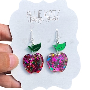 May include: A pair of apple-shaped earrings with a glittery, multi-colored design. Each earring has a green leaf accent and silver-colored ear wires. The earrings are displayed on a white card with the text "Allie Katz Design Studio."