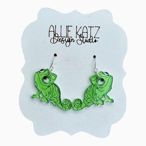 May include: A pair of green chameleon earrings with black outlines and silver hooks. The earrings are displayed on a white, cloud-shaped card with the text "ALLIE KATZ Design Studio" in black script.