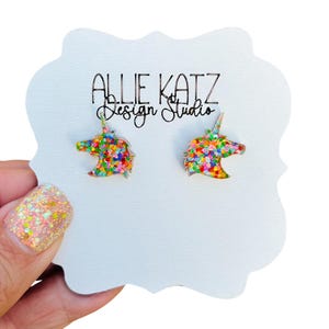 May include: A pair of unicorn stud earrings with a rainbow confetti design. The earrings are on a white card with the text "Allie Katz Design Studio".