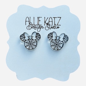 May include: A pair of clear acrylic stud earrings shaped like Minnie Mouse heads, each featuring a black spiderweb design within the ears and bow. The earrings are displayed on a light blue card with the text "Allie Katz Design Studio".
