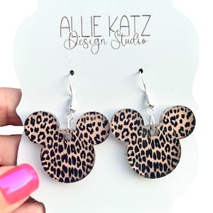 May include: A pair of dangle earrings featuring a Mickey Mouse head shape with a leopard print design. The earrings are attached to silver-colored fish hook ear wires. The card behind the earrings reads "ALLIE KATZ Design Studio."