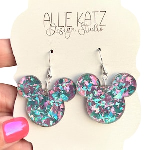 May include: A pair of iridescent Mickey Mouse-shaped earrings. The earrings are filled with colorful glitter pieces in shades of teal, purple, and pink. The text "ALLIE KATZ Design Studio" is printed on the backing card.