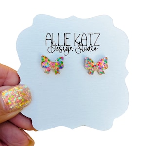 May include: A pair of rainbow glitter bow earrings on a white card with the text "Allie Katz Design Studio".