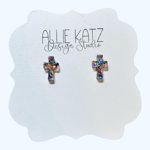May include: A pair of cross-shaped stud earrings. The earrings are adorned with colorful glitter, set against a clear background. The earrings are displayed on a white, cloud-shaped card with the text "Allie Katz Design Studio".
