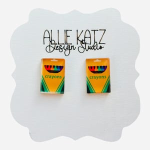 May include: A pair of earrings featuring miniature crayon box designs. The earrings are yellow with green and black accents, and the word "crayons" is printed on the front. The earrings are displayed on a white, decorative card with the text "Allie Katz Design Studio".