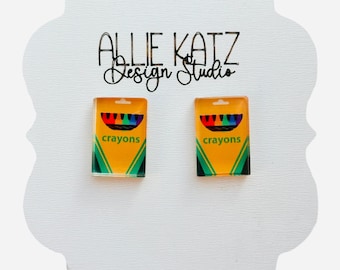 Może przedstawiać: Kolczyki z miniaturowymi wzorami pudełek kredek. Kolczyki są żółte z zielonymi i czarnymi akcentami, a na przodzie nadrukowano słowo "crayons". Prezentowane są na białej karcie z napisem "Allie Katz Design Studio".