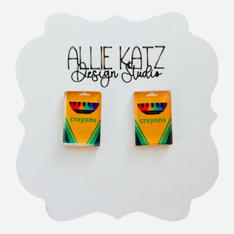 Puede incluir: Pendientes con dise&ntilde;os de cajas de l&aacute;pices de colores en miniatura. Los pendientes son amarillos con detalles verdes y negros, y la palabra "crayons" est&aacute; impresa en la parte delantera. Se presentan en una tarjeta blanca con el texto "Allie Katz Design Studio".