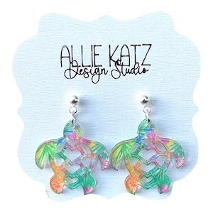 May include: A pair of colorful turtle-shaped earrings with a tropical design. The earrings are made of a clear material with a heart-shaped cutout in the center. The earrings have a silver post and are set against a light blue background with the text "ALLIE KATZ Design Studio".