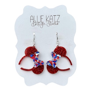 May include: A pair of red glitter earrings with a butterfly design. The butterflies are made of red, white, and blue glitter. The earrings are on a white card with the text "ALLIE KATZ Design Studio".