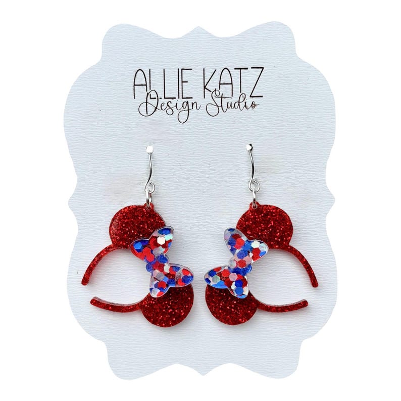 May include: A pair of red glitter earrings with a butterfly design. The butterflies are made of red, white, and blue glitter. The earrings are on a white card with the text "ALLIE KATZ Design Studio".