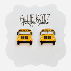 May include: Two yellow school bus earrings with black detailing and the words "SCHOOL BUS" above the windshield. The earrings are on a white background with the text "ALLIE KATZ Design Studio".