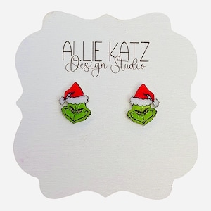 May include: A pair of green Grinch face stud earrings, each wearing a red Santa hat with white trim. The earrings are displayed on a white card with the text "ALLIE KATZ Design Studio" in black script.