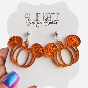 May include: A pair of orange glitter pumpkin earrings in the shape of Mickey Mouse ears. The earrings are hanging from silver posts and are displayed on a white card with the text "Allie Katz Design Studio".