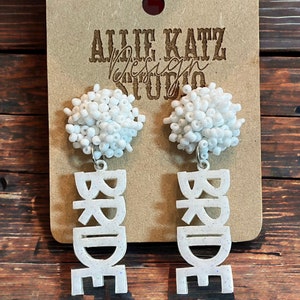 May include: White beaded earrings with the word "BRIDE" spelled out in white glitter. The earrings are on a brown background with a white card that says "Allie Katz Design Studio".