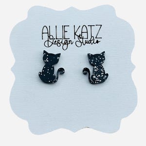 May include: A pair of black cat-shaped stud earrings with white glitter accents. The earrings are displayed on a light blue, scalloped card with the text "Allie Katz Design Studio" in black script.