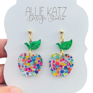 May include: A pair of apple-shaped earrings with a green leaf charm. The apple is clear and filled with colorful confetti.