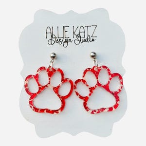 May include: A pair of red and white paw print earrings. The earrings are made of a clear material with red spots and are attached to silver-colored posts. The earrings are displayed on a white card with the text "Allie Katz Design Studio".