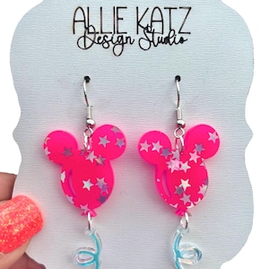 May include: A pair of bright pink Mickey Mouse-shaped earrings with silver star accents. The earrings have silver hooks and clear, iridescent balloon string details. The earrings are displayed on a white card with the text "Allie Katz Design Studio".