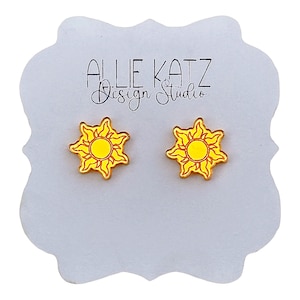 May include: A pair of bright yellow sun-shaped stud earrings with orange outlines, displayed on a white card featuring the text ALLIE KATZ Design Studio. The earrings have a central circle surrounded by six stylized, flame-like sun rays.