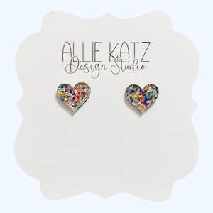 May include: A pair of heart-shaped stud earrings with a glittery, multicolored design. The earrings are displayed on a white card with the text "ALLIE KATZ Design Studio" in black script.