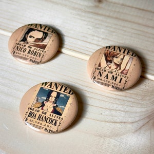 May include: Three round, brown, button-shaped pins with a wanted poster design. Each pin features a different character from the anime One Piece. The posters feature the characters Nico Robin, Boa Hancock, and Nami. The posters have the text "WANTED" at the top, "DEAD OR ALIVE" in the middle, and the character's name and bounty below. The posters are surrounded by a brown border.