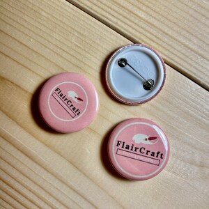 May include: Two pink button badges with a white logo that says "FlairCraft" and a stylised image of a crab. One badge has the text "Clothing Memories" below the logo. The other badge has a plain back with a pin back.