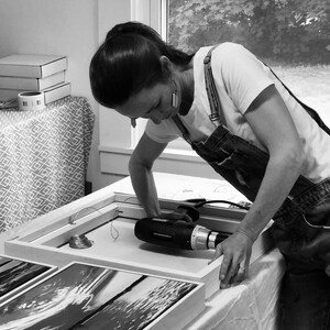 May include: A person using a power drill to assemble a framed artwork. The artwork is a black and white photograph. The person is wearing a white t-shirt and denim overalls. Other framed artworks are visible.