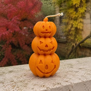 May include: A stack of three orange pumpkin-shaped candles with carved jack-o'-lantern faces. The top pumpkin is the smallest, the middle is medium, and the bottom is the largest. A single light-colored wick extends from the top of the stack.