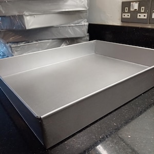 Puede incluir: Una pila de moldes para hornear rectangulares de color plateado. Un molde está en primer plano, con forma cuadrada y lados rectos. Los moldes parecen ser de metal y están apilados sobre una superficie oscura.