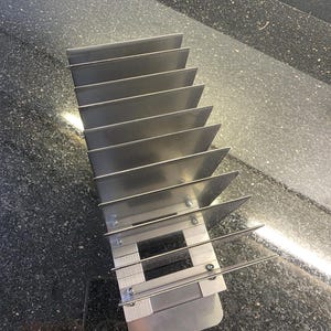 Peut inclure: Un support en métal argenté composé d'une série de séparateurs verticaux parallèles fixés sur une base centrale. La structure est formée de plusieurs plaques rectangulaires fines alignées, maintenues par des vis visibles sur le cadre.