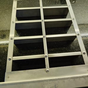 Puede incluir: Un cortador de dulce de azúcar rectangular de acero inoxidable con una rejilla de divisores verticales y horizontales asegurados con tornillos metálicos. La herramienta incluye un mango ancho en la parte superior y una base con la palabra Fudge grabada.