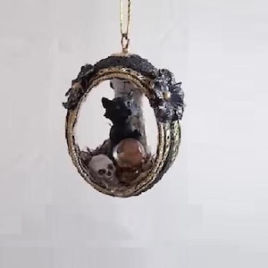 May include: A black cat ornament with a gold and black frame, holding a skull and a red gem. The ornament is hanging from a gold string.