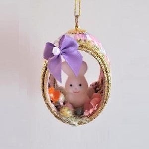 May include: A pink and gold Easter egg ornament with a purple bow and a small white bunny inside. The bunny is sitting on a bed of pink and white flowers and there are gold and orange accents.